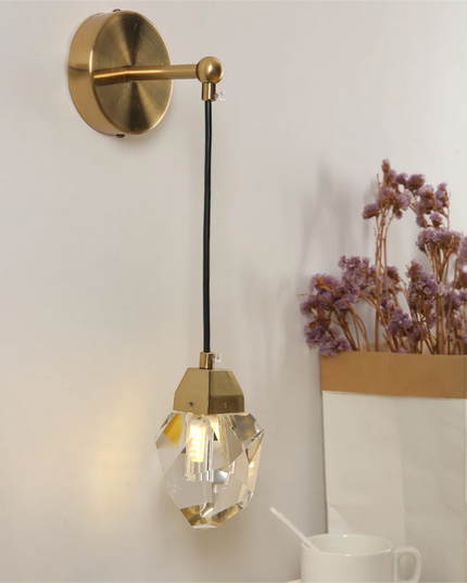 Gold wall-mounted lamp with crystal shade on a white wall, next to a vase of dried flowers.