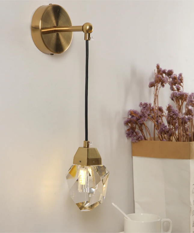 Gold wall-mounted lamp with crystal shade on a white wall, next to a vase of dried flowers.