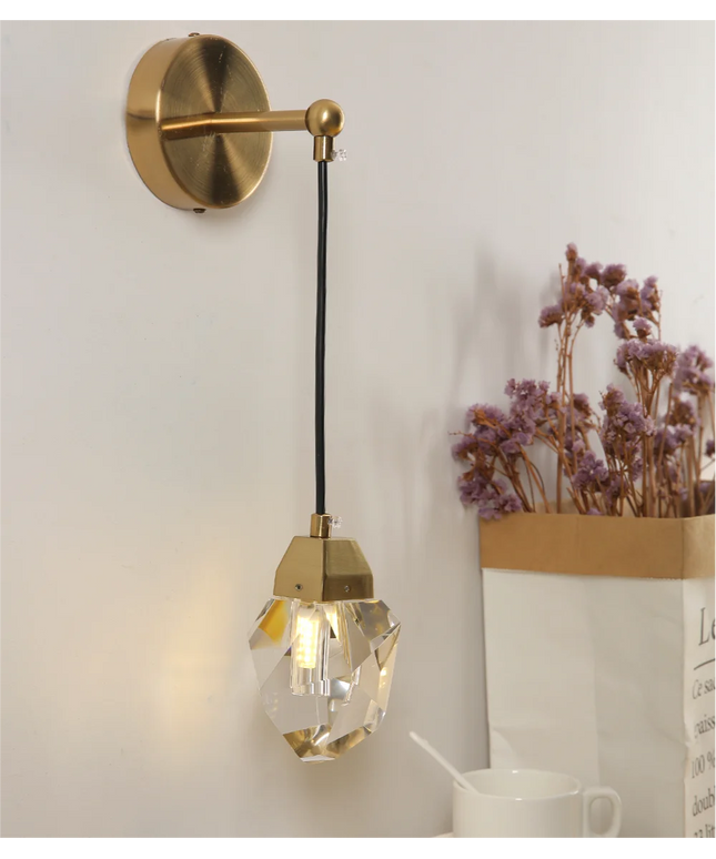 Gold wall-mounted lamp with crystal shade on a white wall, next to a vase of dried flowers.