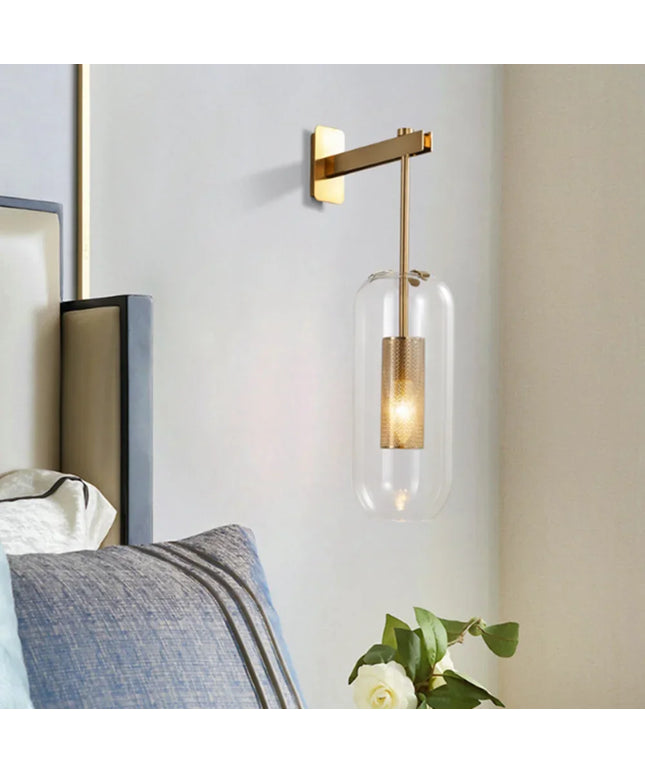 Modern wall sconce with gold finish and clear glass shade in a bedroom setting.